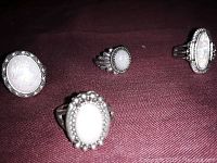 Four silver tone rings with white oval and round stones on a burgundy cloth background, showing the ring details and condition.