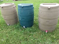 Three heavy plastic rain barrels in grass, two tan and one green, showing general wear and outdoor use.