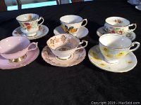 Six varied signed and numbered tea cups with matching saucers displayed on a black surface, showing different floral and decorative patterns.