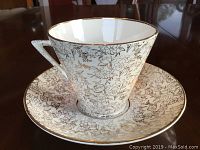 Tea cup and saucer set showing gold floral inlay and triangular handle with gold trim, photographed against a brown reflective surface.