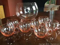 Photo of 8-inch footed light pink crystal punch bowl and 7 of the 11 matching cups arranged around it on a wooden table.