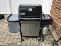 Full frontal of the Weber Genesis Silver gas grill showing side tables, cabinet, wheels, control panel, and attached propane tank.