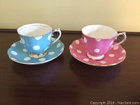 Two tea cup and saucer sets, one blue with white polka dots and one pink with white polka dots, both with gold trim, displayed on a wood surface.