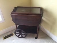 Wooden tea trolley with glass tray top, side and wheels shown from angled view near window and vent on floor.