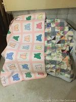 Two quilts side by side. Left quilt has butterfly applique design in pastels with pink border. Right quilt is patchwork of various patterned fabric triangles in soft colors.