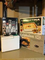 Lot consisting of Braun hand blender in box, Lagostina roasting pan box, and Calphalon contemporary nonstick pan box stacked on top of a white surface.