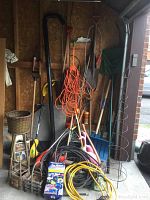 Photo showing multiple long-handled garden tools hanging and leaning against a plywood wall, including rakes, shovels, coiled extension cords, hoses, and other items piled at the base.