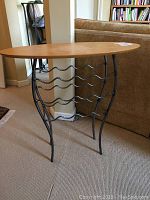 Side angle view showing the wooden oval table top and black metal wine rack base with five wavy metal rails.