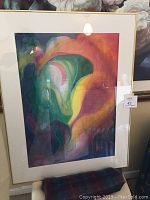 Framed abstract watercolour painting leaning against a wall, showing colorful organic shapes in green, purple, yellow, and orange with a gold frame and white mat.