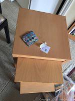 Three light wood nesting tables stacked together with a colorful square coaster on top. Smallest table shows a chip on one corner.