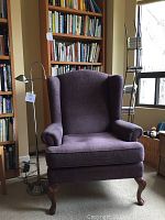 Front view of plum wing back chair next to metal reading lamp and wooden bookshelf