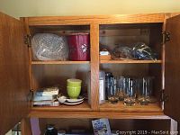 Open wooden cabinet with various glasses, green cup and saucer, paper napkins, and other items visible on shelves.