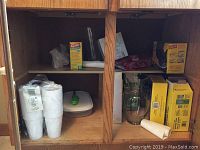 Wooden cabinet with clear glass vase, boxes of Glad garbage bags, lamp oil gift set box, white disposable plastic cups, green and white plastic container.