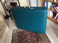 Closed portable massage table in teal vinyl upholstery resting on carpet, standing vertically with handle visible.