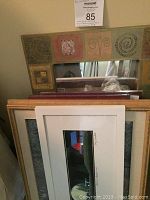 Wide view of brown, gold, and decorative frame hand-painted mirror with other framed items stacked underneath including framed prints and frames.