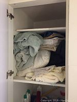 Open white cabinet with shelves holding folded towels, bed sheets, pillows and a shower curtain.