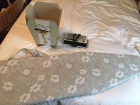 Photo showing ironing board cover, decorative waste basket with bird painting, and clock radio on bed.