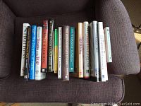 Fourteen self-help and business books laid out on a purple cushioned chair, titles clearly readable, covering topics including leadership, team management, coaching, and mindfulness.