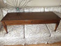Full view of the rectangular brown wood coffee table with tapered legs, placed on a floral patterned couch for scale.