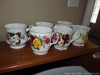 Six bone china coffee mugs with different floral patterns displayed on a wooden surface, showing variety in flower colors and design.