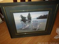 Framed print showing two white Adirondack chairs on a dock overlooking a calm lake with an island of evergreen trees; image shows overall condition and framing.