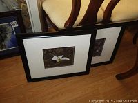 Two black framed and matted nature photographs leaning against chairs, one with a white flower on natural ground, the other partially visible behind it.