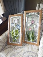 Two wood framed stained glass window panels displayed on a chair with a blue backrest and patterned cushion. Both panels have floral designs and clear textured glass backgrounds.