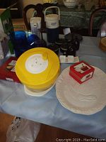 Image of kitchen lot showing yellow and white salad spinner at center, Coleman Polylite beverage cooler jug behind it, blue acrylic bowls, white ceramic platter with embossed vegetables on the right, small ceramic dip bakers and salt and pepper shakers, mugs, and boxes.