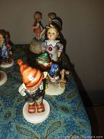 Various angles of four Hummel figurines and one Hummel music box arranged on a patterned cloth surface.