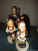Four Goebel Hummel figurines displayed together on fabric surface. Three boys in traditional attire, one holding book, others with accessories.