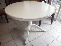 White round kitchen table showing top and part of base with four legs, placed on tiled floor with surrounding chairs.