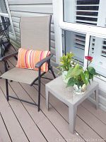Outdoor chair with mesh seat and backrest, black metal frame, striped pillow on chair, small gray plastic table with two potted plants on wooden deck near house window