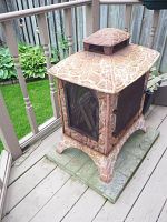 Square metal fireplace with mesh screen door and textured stone-like pattern on metal panels, resting on rectangular fireproof base on a wooden deck.