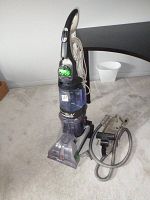 Front view of the Hoover Steam Vac Dual carpet cleaner with hose attachment placed on carpet. Clear water tank with green brushes visible.