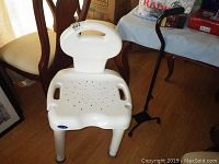 Photo of white Invacare bath chair with backrest and drainage holes, next to black adjustable cane with ergonomic handle, both placed on wooden floor indoors.