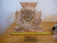 Front view of a pinkish onyx mantel clock with round clock face, Roman numerals, and decorative carved body on a flat base.