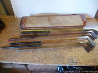 Overview of vintage canvas golf bag lying flat beside six wood shaft golf irons arranged horizontally on a table.