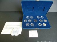 Full view of the blue velvet-lined display case holding 13 commemorative Canadian coins in clear capsules, accompanied by informational papers.