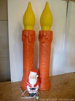 Photo showing two large orange candle blow molds with yellow flame tops and a small Santa blow mold in front. The candles have 'NOEL' embossed vertically.