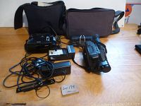 Two video cameras — Hitachi E31A and Sanyo Zeema Fuzzy Logic — displayed with respective chargers, cables, and carrying bags on wooden surface.