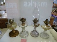 Four vintage oil lanterns displayed side by side on white surface. One lantern has a glass chimney; all have metal burners. Lanterns are between 10 and 11 inches tall.
