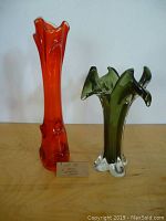 Two hand blown glass vases, one red tall and one green shorter, displayed on a wooden surface with white background. Both show flared ruffled top edges and thick bases.