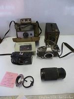 Four cameras displayed with the Soligor zoom lens and two metal mount rings on white surface. Cameras include Olympus OM-10 (missing lens), vintage Polaroid 220 with flash, Brownie Target Six-16 box camera with side handle, and lexus 35mm camera with strap. Lens and rings placed near cameras.