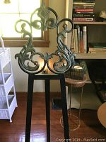 Black metal decorative easel with ornate scrollwork visible from front, standing on wood floor near window and shelving.