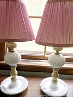 Photo of two boudoir lamps with white milk-glass bases and pink pleated fabric shades on a wooden surface near a window.