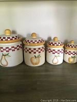 Four ceramic glass canisters with fruit designs, showing lids and checkered red accent bands, arranged from large to small.