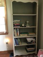 Full view of pale green bookshelf with books on four shelves, two stuffed teddy bears on the second shelf, and a decorative storage box on the fourth shelf.