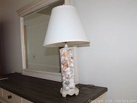 Table lamp with clear cylindrical base filled with an assortment of white and beige seashells, topped by a white conical lampshade, sitting on a wooden surface in front of a mirror.