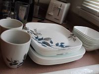 Stack of square dinner plates, smaller plates, bowls, and two white coffee cups on a kitchen counter.