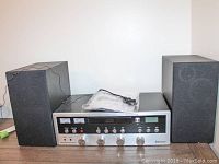 Front view of stereo main unit with two speakers on either side, manual on top visible. Silver face with control knobs and small display.